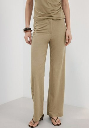 Osoba ubrana w luźne beżowe spodnie, dopasowany top, ciemne szerokie bransolety na lewym nadgarstku oraz ciemne sandały na koturnie z cienkimi paskami.