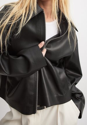 Blonde person wearing a partially zipped black leather jacket over a white shirt, with white pants and hands tucked in pockets.