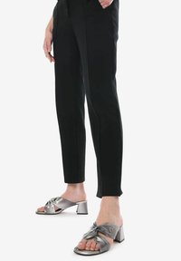 Mule aperte in metallo argento con un design annodato sulla parte superiore, tacco spesso e una texture liscia. Abbinate a pantaloni neri.