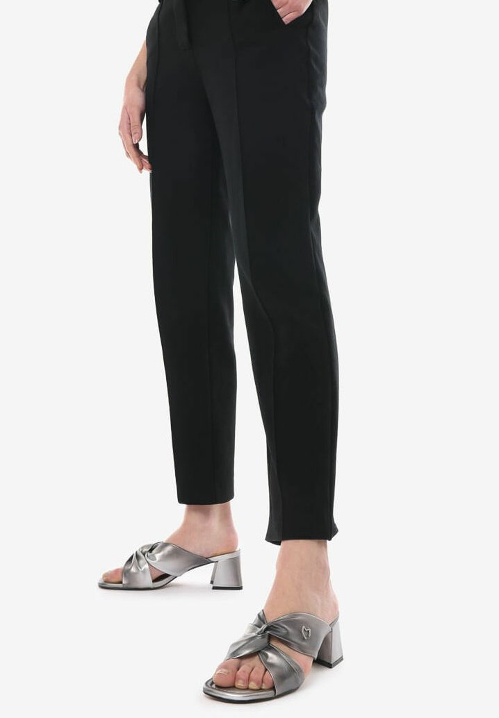 Mule aperte in metallo argento con un design annodato sulla parte superiore, tacco spesso e una texture liscia. Abbinate a pantaloni neri.