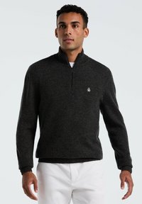 Mann trägt einen dunkelgrauen Pullover mit Viertelreißverschluss und weiße Hose, steht vor einem schlichten hellgrauen Hintergrund.