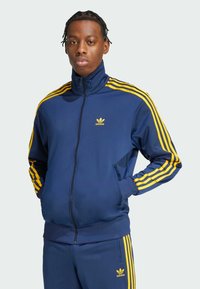 Tamno plavi trenirku s žutim trostrukim akcentima na rukavima, puni patentni zatvarač i mali žuti Adidas logo na prsima. Glatka tekstura tkanine.