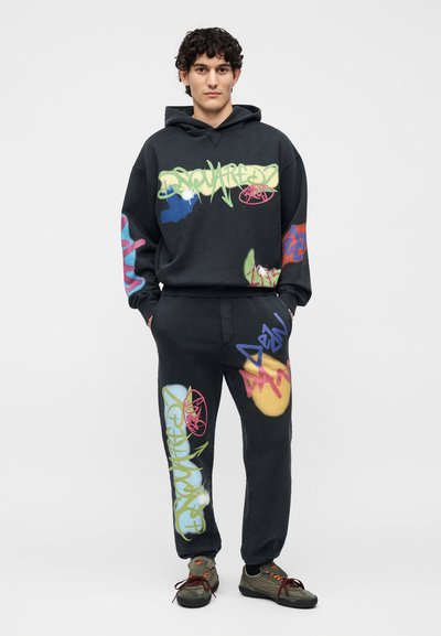Νέος άνδρας που φοράει μαύρο φούτερ με κουκούλα και φόρμες με πολύχρωμες εκτυπώσεις σε στυλ γκράφιτι, σε συνδυασμό με πράσινα και μαύρα αθλητικά παπούτσια.