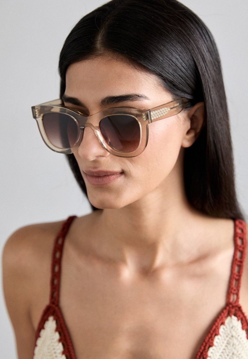 Transparent acetate sunglasses with gradient brown lenses; square frame and textured gold accents on arms; worn by a model with dark hair.