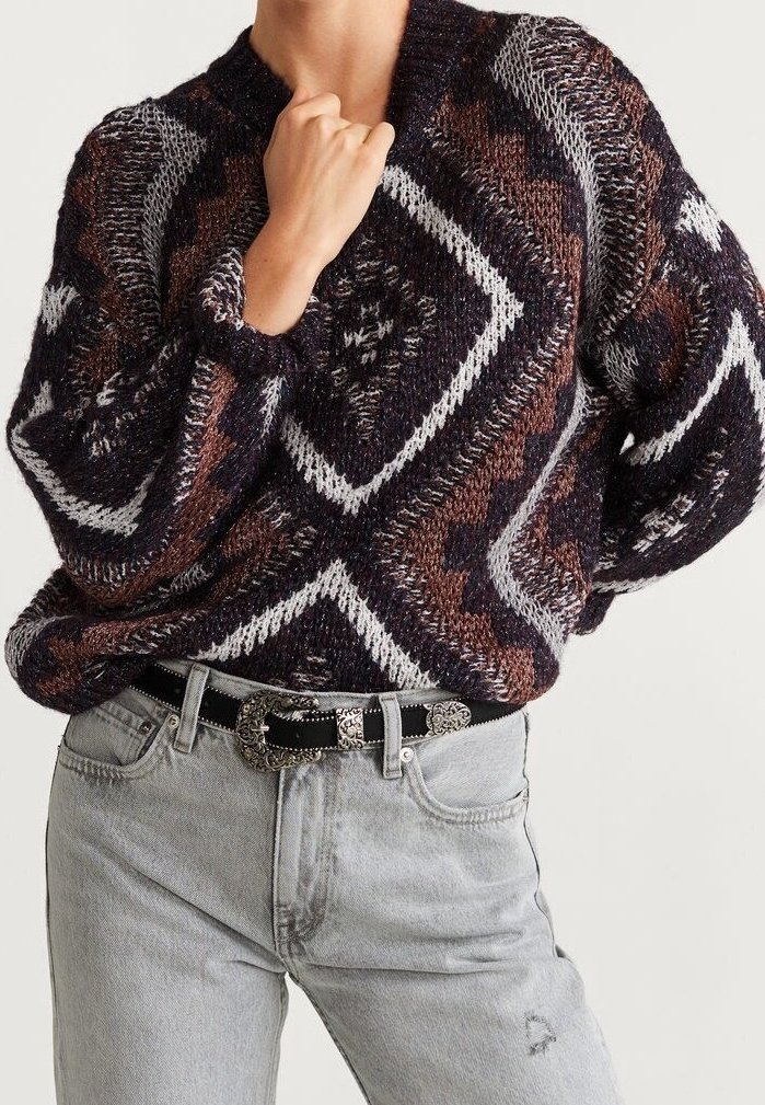 Person wearing a patterned knit sweater in dark brown, black, and white, paired with light gray jeans and a black belt with an ornate silver buckle.