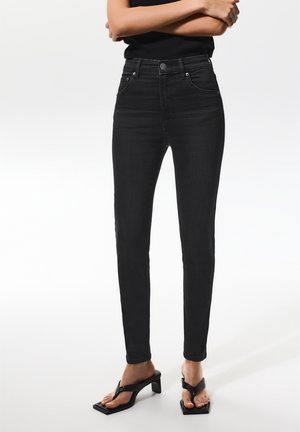 Vrouw draagt hoge taille zwarte skinny jeans gecombineerd met zwarte sandalen met hakken, staand met gekruiste armen tegen een witte achtergrond.