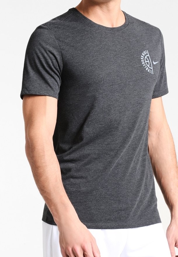 Mężczyzna w ciemnoszarym podkoszulku do koszykówki Nike i białych szortach, stojący z rękami swobodnie opuszczonymi wzdłuż ciała.