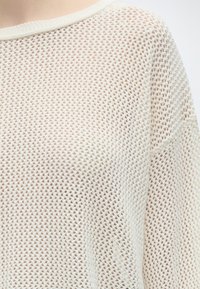 Gros plan d'un pull blanc à maille lâche montrant son motif texturé en tissage ouvert et son encolure arrondie.