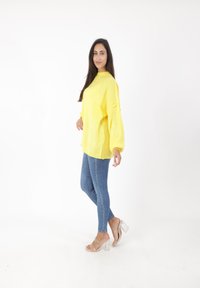 Maglione giallo oversize a maglia con maniche a palloncino, abbinato a jeans blu aderenti e sandali trasparenti con tacco. Sfondo bianco.
