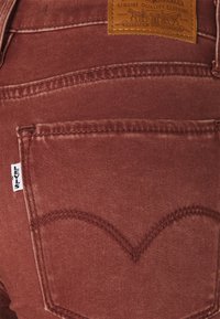 Jeany v rezavé barvě s měkkou texturou, se zvláštním zakřiveným designem zadních kapes a hnědou koženou nášivkou na pásku.