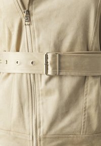 Giacca beige con zip, dal tessuto liscio e morbido, dotata di una larga cintura con fibbia metallica e tasche laterali.