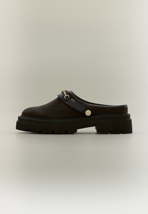 Chaussure à enfiler marron foncé avec semelle épaisse et robuste, boucle dorée décorative et petit talon arrière sur fond uni.