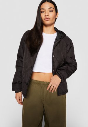 Kvinde med langt mørkt hår iført en sort quiltet jakke over en hvid cropped top og olivengrønne bukser mod en ensfarvet grå baggrund.