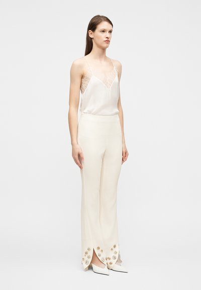 Γυναίκα που στέκεται φορώντας λευκό κορμάκι με κρέμ δαντέλα και ψηλόμεσα ελεφαντόδοντα παντελόνια με διακοσμητικά λουλούδια κοντά στις σχισμές, με λευκά μυτερά παπούτσια.