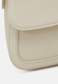 Nærbillede af hjørnet af en beige pebret læderhandtaske med syet flapdetalje på hvid baggrund.