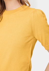 Žlutý krátký rukávový top s kulatým výstřihem a nařasenými rameny. Vyrobeno z měkkého, lehkého materiálu s hladkou texturou.