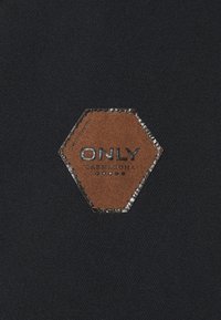 Tessuto nero con superficie strutturata e toppa esagonale in pelle marrone con il marchio "ONLY CARMAKOMA" in rilievo.