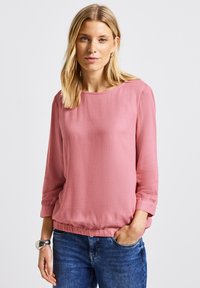 Blusa rosa con finitura testurizzata, maniche a tre quarti e vestibilità morbida. Collo rotondo e orlo inferiore elasticizzato.