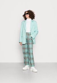 Blazer verde menta claro, camisola de gola alta branca, calças largas de xadrez em teal e castanho, combinadas com sapatilhas brancas de plataforma. Óculos de sol pretos usados.
