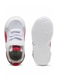 Baskets blanches en mesh avec des accents rouges, une languette rembourrée et une sangle Velcro. La semelle présente un motif en caoutchouc texturé blanc.
