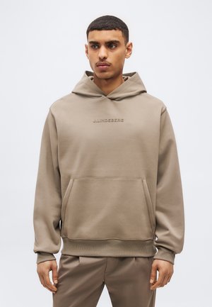Mladi moški, oblečen v bež kapucar s sprednjim žepom in subtilnim logotipom, stoji pred enotnim svetlim ozadjem in rahlo gleda proti desni.