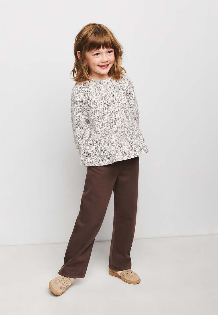Jeune fille aux cheveux bruns mi-longs portant un haut blanc à manches longues à motifs, un pantalon marron et des chaussures beiges, debout devant un fond uni.