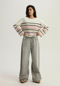 Przytulny kremowy sweter z wielokolorowymi poziomymi paskami, w połączeniu z jasnoszarymi spodniami o szerokich nogawkach i brązowymi butami. Swobodny, luźny krój.