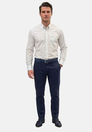 Hombre de pie que lleva una camisa blanca de manga larga con estampado, pantalones azul marino, un cinturón trenzado azul marino y zapatos negros.