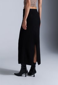 Zwarte gebreide maxi-rok met een zijsplits, zachte textuur, hoge tailleband, gedragen met zwarte haklaarzen, en toont een getailleerd ontwerp.