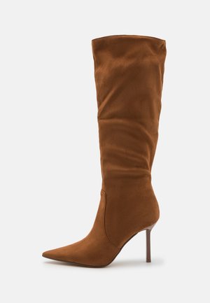 Bottes en daim marron montantes avec un bout pointu et un talon aiguille, présentant un design supérieur en mode slouch contre un fond blanc.