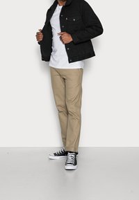 Černá džínsová bunda s sherpa podšívkou, bílý tričko, béžové kalhoty a černo-bílé tenisky. Čistý, ležérní design outfitu.