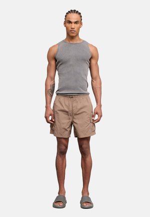 Uomo con capelli intrecciati che indossa una maglietta grigia senza maniche, pantaloncini beige con coulisse e sandali grigi, in piedi rivolto in avanti.