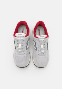 Pár světle šedých tenisek New Balance 574 s červeným vnitřním podšívkou, bílými tkaničkami a síťovinou na vrchu, nahlédnuto zhora na bílém pozadí.