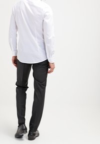 Chemise blanche à manches longues avec poignets boutonnés et pantalon habillé noir, présentant une coupe droite et un tissu lisse. Des chaussures noires complètent la tenue.