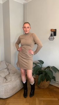 Bež obleka z dolgimi rokavi z rebrastim vzorcem in visokim izrezom, kombinirana z črnimi škornji do kolen. V prostoru je teksturirana bež sedežna garnitura in zelena rastlina.