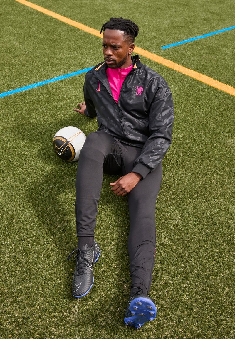 Giacca sportiva nera con accenti rosa, pantaloni neri aderenti e scarpe da calcio. Palla da calcio bianca con dettagli neri e dorati su erba verde.