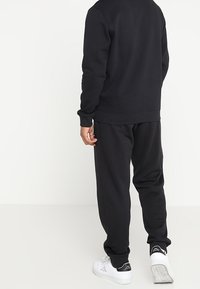 Person som bär svart sweatshirt och mjukisbyxor, vita sneakers med logotyp, står mot en enkel vit bakgrund, vänd bort.