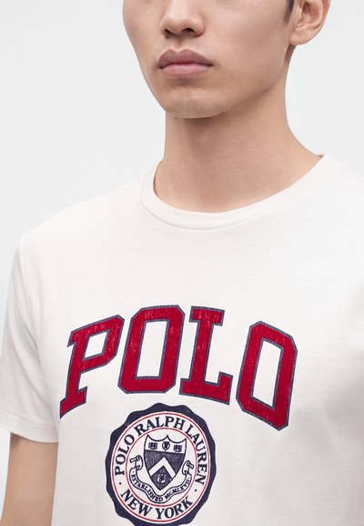 Νέος άνδρας που φοράει λευκό T-shirt με στρογγυλή λαιμόκοψη, μεγάλο κόκκινο κείμενο "POLO" και έναν κυκλικό λογότυπο Polo Ralph Lauren New York από κάτω.