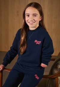 Conjunto de sudadera y pantalones de chándal en azul marino, hecho de algodón. Presenta el logo rojo de "Tommy" en el pecho y el muslo. Puños de canalé y corte relajado.