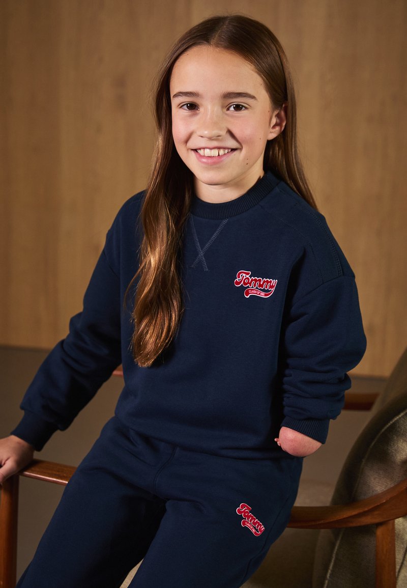 Conjunto de sudadera y pantalones de chándal en azul marino, hecho de algodón. Presenta el logo rojo de "Tommy" en el pecho y el muslo. Puños de canalé y corte relajado.