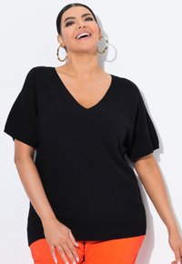 Černý pletený top ve tvaru V s krátkými rukávy, spárovaný s jasně oranžovými kalhotami. Tkanina vypadá měkce a má strukturu, což nabízí uvolněný střih.