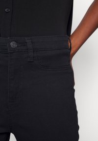Gros plan d'une personne portant un jean noir taille haute avec un bouton, la main reposant près de la poche, sur un fond blanc.