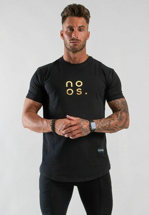 Muskelös man med stylat hår som bär en svart noos t-shirt, svarta leggings, tatuerad höger arm, smartklocka och svart pärlarmband, står med händerna knäppta.