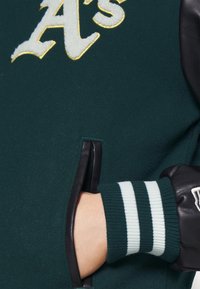 Veste en polaire verte avec manches en cuir noir, arborant des lettrages blancs et un poignet rayé vert et blanc. Poche latérale visible.