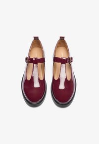 Chaussures en cuir verni bordeaux avec semelles noires, bride T à la cheville, bout rond et texture lisse. Dotées d'une petite boucle et d'un intérieur beige.