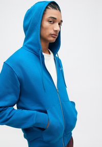 Sudadera azul con cremallera, con un bolsillo frontal y capucha con cordón, que presenta una textura suave y puños acanalados, llevada sobre una camiseta blanca.