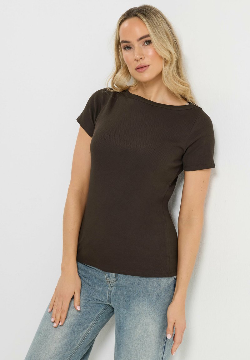 Femme blonde portant un haut noir ajusté à manches courtes et un jean bleu clair, debout contre un mur blanc avec une main posée sur sa cuisse.