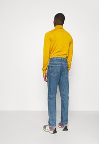 Suéter amarillo de cuello alto, pantalones vaqueros azules con bolsillos traseros y zapatillas multicolores con detalles en púrpura y verde, vistos desde atrás.