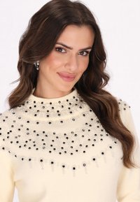 Pulover de culoare crem, cu gât înalt, împodobit cu mărgele negre și albe aranjate într-un model, având o textură moale și un design ajustat.