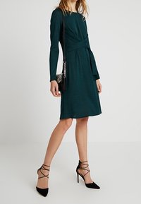 Tmavě modré šaty s dlouhým rukávem, zavinovací design a zavázaným pasem. Nosené s černou crossbody taškou a černými sandály na vysokém podpatku. Hladká textilie.